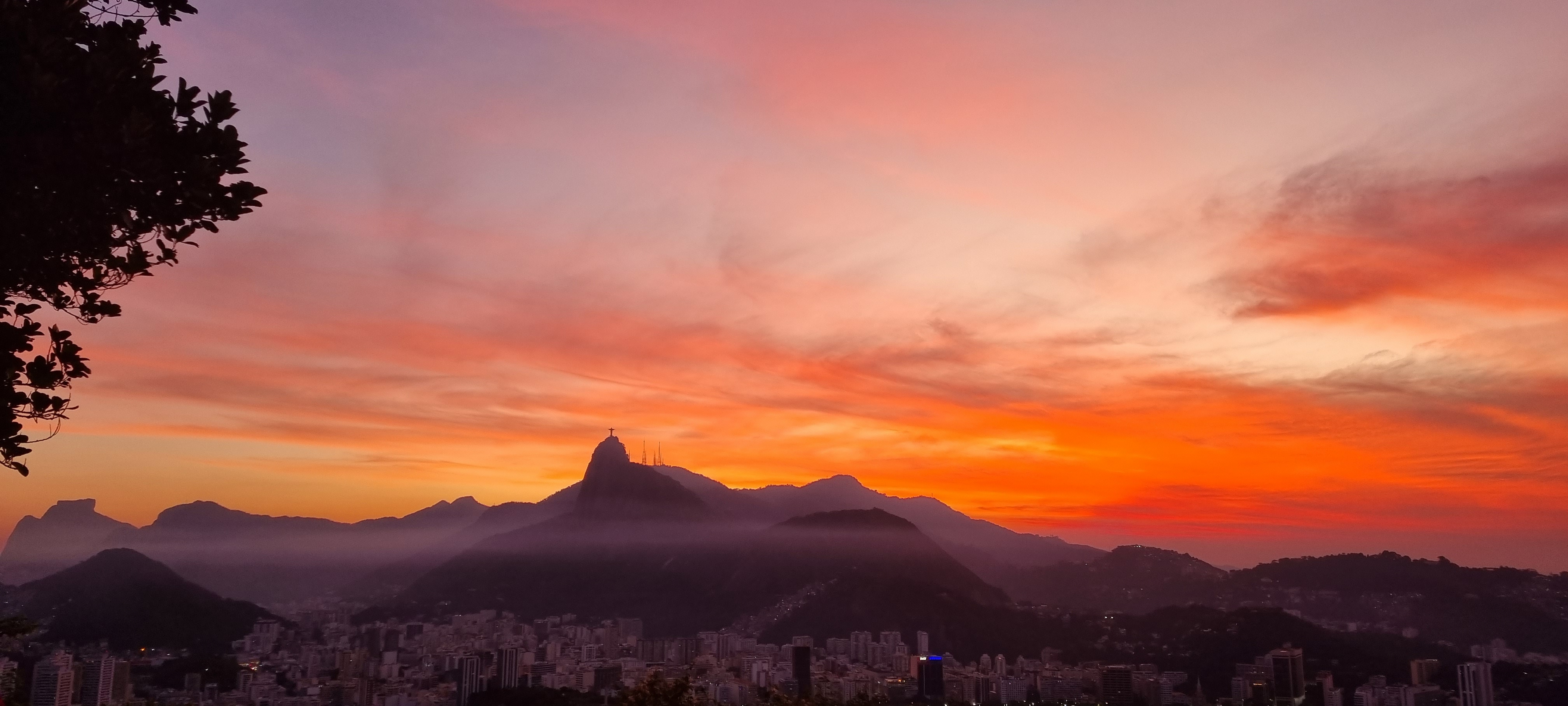 View of Rio de Janeiro from the top of the Sugarloaf Mountain. It is sunset, so the sky has a lot of red, orange and yellow tones. Christ the Redeemer can be seen in the middle of the picture.
