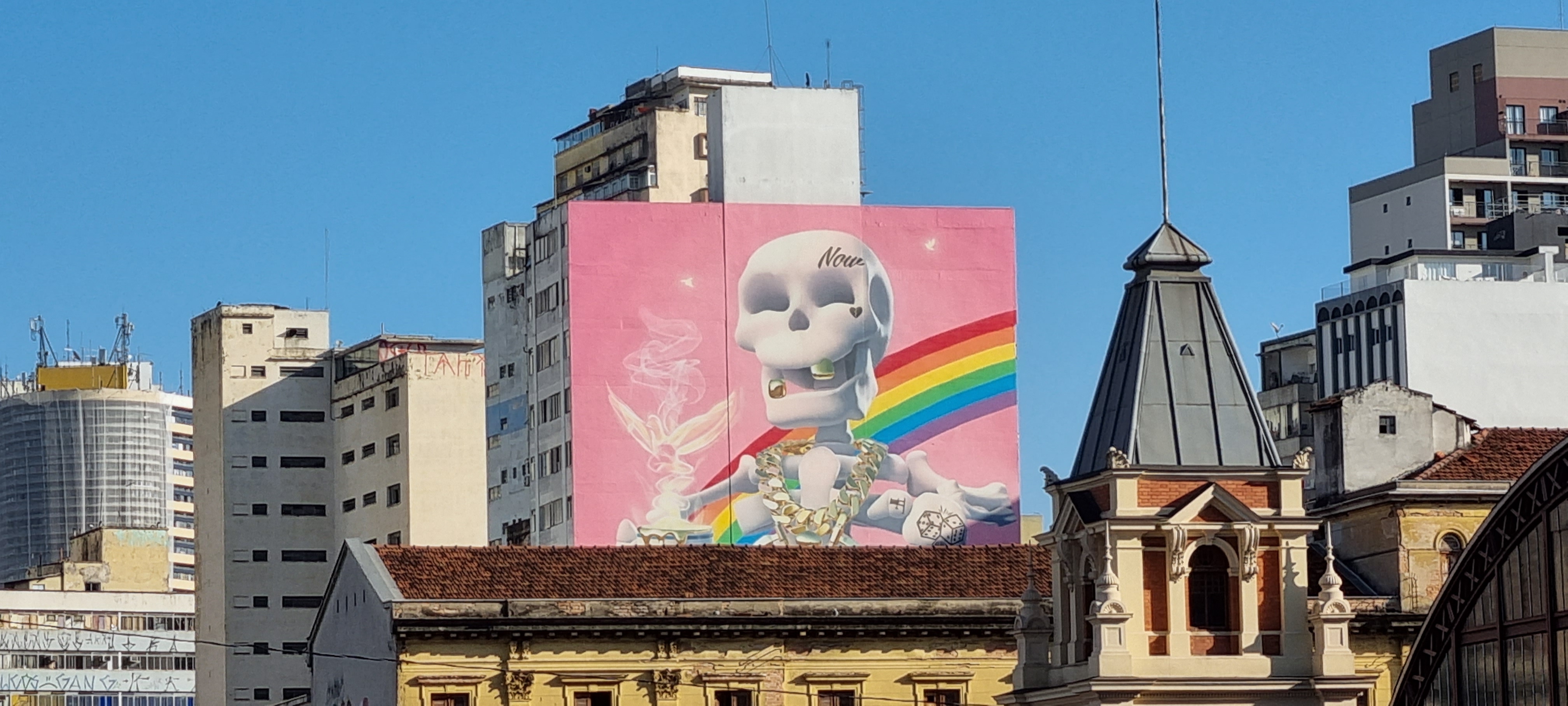 Picture of an urban landscape in which in one of the buildings you can see a mural of a funky skeleton with a rainbow in the background.